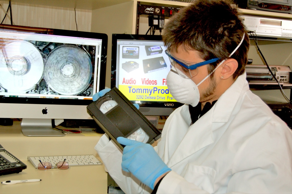 Technician in a mask and gloves inspecting a VHS tape
